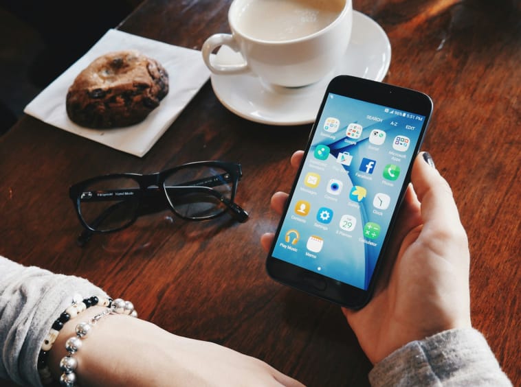person holding turned-on smartphone sitting at table with filled cup with saucer